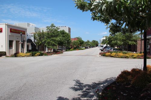A quiet street lined with trees and colorful flowers, featuring buildings and a clear blue sky.
