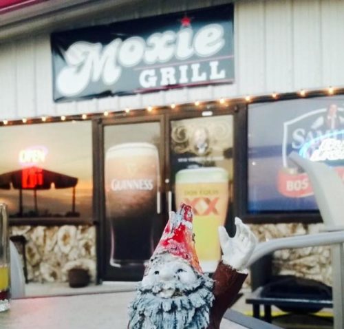 A small gnome figurine with a red hat stands on a table in front of the Moxie Grill, featuring a bar sign in the background.