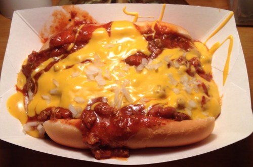 A hot dog topped with chili, melted cheese, and diced onions in a paper tray.