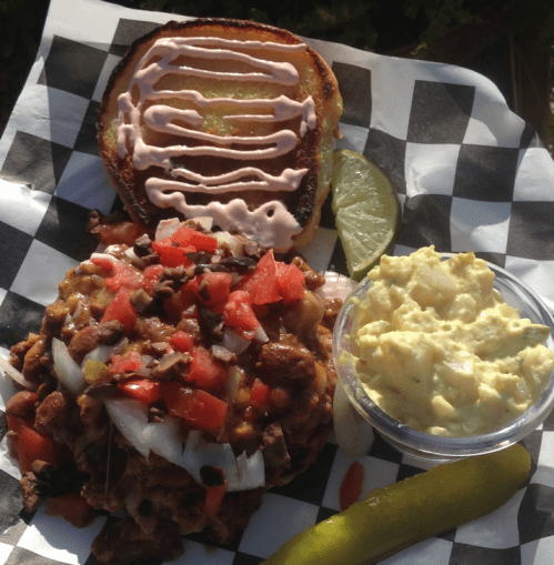 A plate featuring a burger with toppings, a side of egg salad, a pickle, and a lime wedge on a checkered paper.