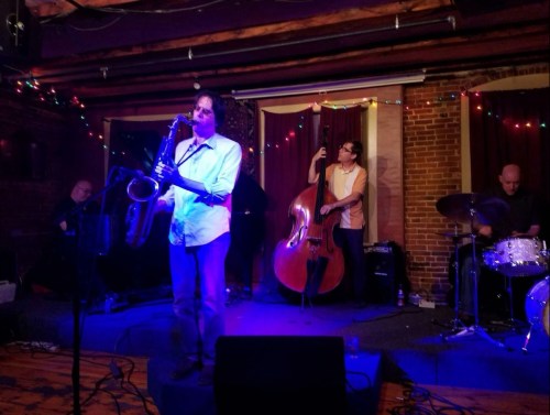 A jazz band performs on stage, featuring a saxophonist, bassist, and drummer, with colorful lights in a cozy venue.
