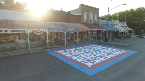 A colorful quilt-patterned mural on a street, with historic buildings and festive decorations in the background.