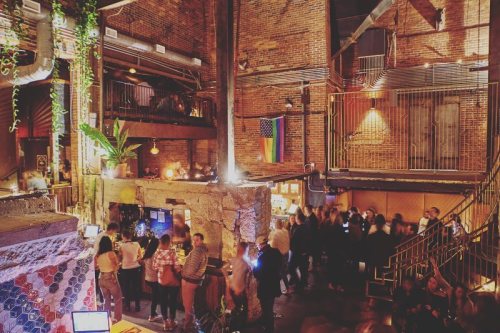 A lively bar scene with exposed brick walls, plants, and a rainbow flag, filled with people enjoying the atmosphere.