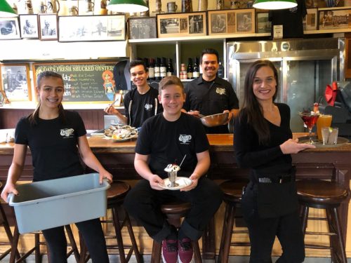 A group of five restaurant staff members pose behind a bar, smiling and holding food and drinks.
