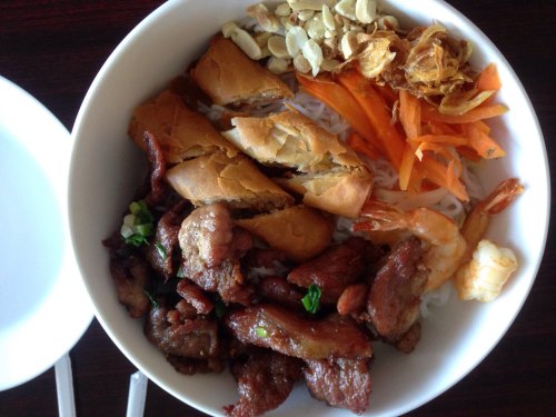 A bowl of vermicelli noodles topped with grilled meat, shrimp, pickled carrots, fried shallots, and crispy spring rolls.