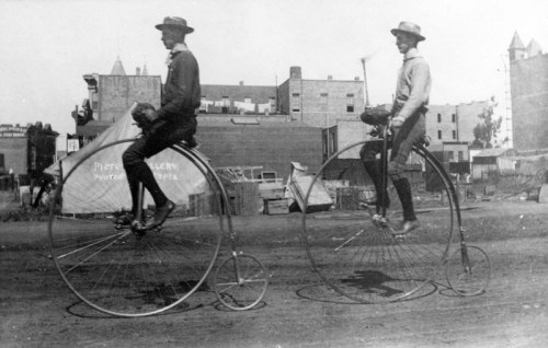 Two men riding penny-farthing bicycles on a dirt street, with buildings and a tent in the background.