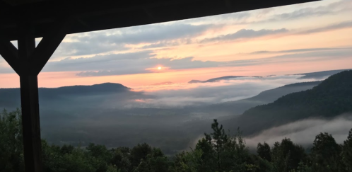 A serene sunrise over misty valleys and hills, viewed from a wooden structure. Soft clouds blanket the landscape.