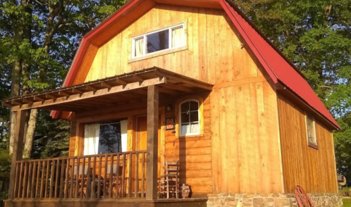 A wooden cabin with a red roof, porch, and large windows, surrounded by trees.