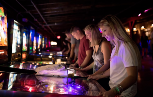 A group of friends playing pinball machines in a dimly lit arcade, focused on the game with colorful lights.