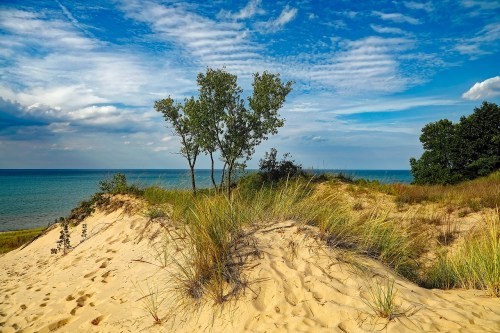 A sandy dune with grass and trees overlooks a calm blue lake under a partly cloudy sky.