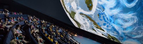Audience seated in a planetarium, gazing up at a large projection of Earth and swirling clouds.