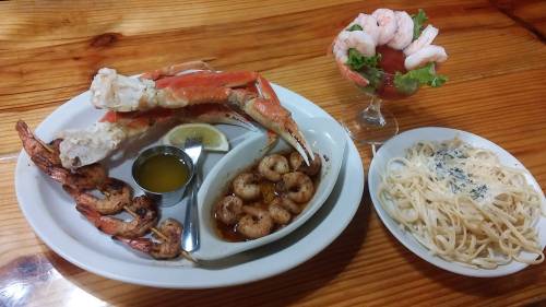 A seafood platter featuring crab legs, shrimp, pasta, and a shrimp cocktail in a glass, served on a wooden table.