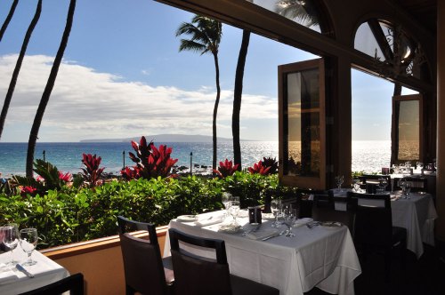 A seaside restaurant with white tablecloths, overlooking the ocean and palm trees, framed by large open windows.