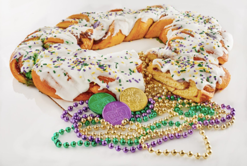 A colorful king cake with icing and sprinkles, surrounded by gold, green, and purple beads and coins.