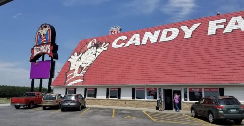 A colorful candy store with a large sign, featuring a cartoon character, and cars parked outside on a sunny day.