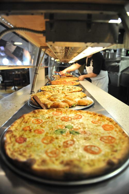 A variety of pizzas displayed on a counter, with a chef preparing food in the background.