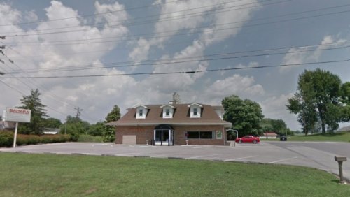 A brick building with a sloped roof, surrounded by grass and trees, under a cloudy sky. A red car is parked nearby.