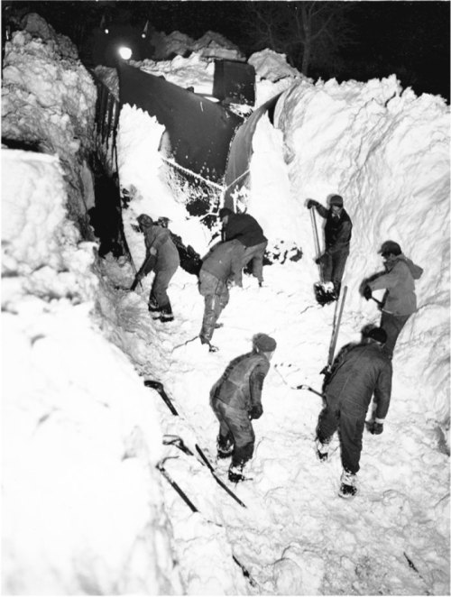 Workers shoveling snow in a deep snowdrift, with a train partially visible in the background at night.