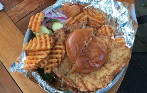 A plate of crispy fried chicken sandwich with waffle fries, pickles, and onion slices, wrapped in foil.