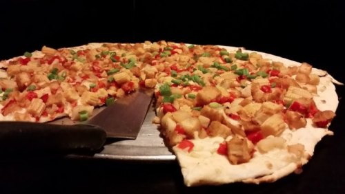 A pizza topped with diced vegetables and crispy pieces, with a slice cut out and a pizza cutter beside it.