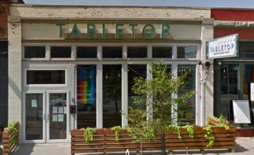Exterior of a shop named "Tabletop," featuring rainbow and American flags, with wooden seating outside.