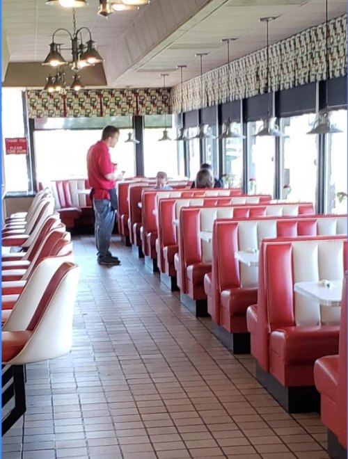 A diner interior with red and white booths, a man standing, and a few customers seated at tables.