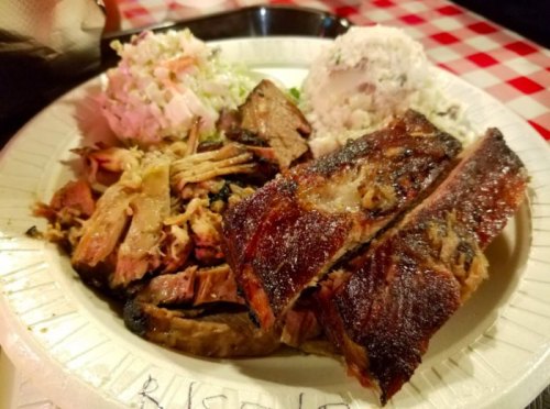 A plate of barbecue featuring sliced brisket, pulled pork, coleslaw, and rice, served on a checkered tablecloth.