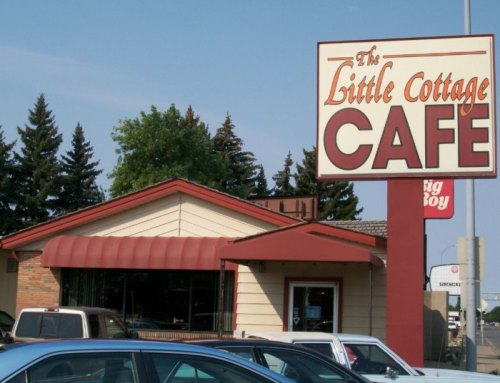 A cozy café with a red awning and a large sign reading "The Little Cottage Café" in a suburban setting.