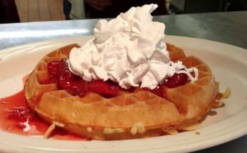 A golden waffle topped with whipped cream and strawberry sauce on a white plate.