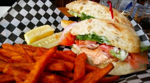 A sandwich with lettuce, tomato, and fish, served with sweet potato fries and a lemon wedge on a checkered plate.