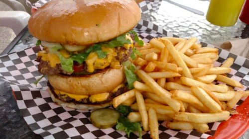 A towering cheeseburger with lettuce and pickles, served with a side of golden French fries on a checkered paper.