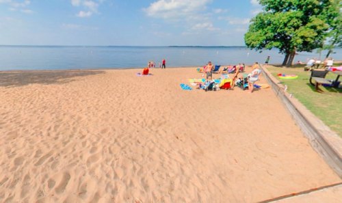 A sandy beach by a calm lake, with people relaxing on towels and colorful beach gear under a clear blue sky.