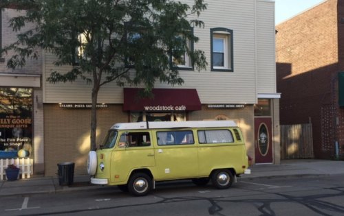 A yellow vintage van parked in front of the Woodstock Café, with trees and buildings in the background.