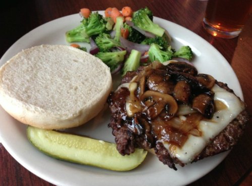 A plate featuring a burger with mushrooms and cheese, a side of mixed vegetables, a pickle, and a bun.