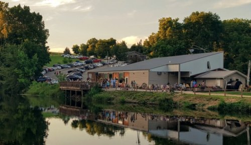 A lakeside building with people outside, surrounded by trees and parked cars on a hill in the background.