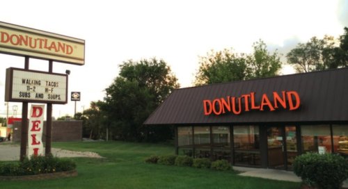 A storefront of Donutland with a sign advertising walking tacos, subs, and soups, surrounded by green grass and trees.