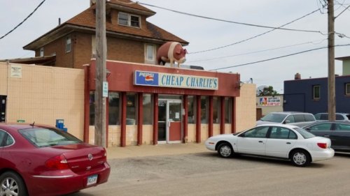 A storefront labeled "Cheap Charlie's" with a pig statue on the roof and parked cars in front.