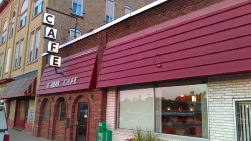 Exterior of a cafe with a red awning and sign, featuring large windows and a brick facade in a small town setting.