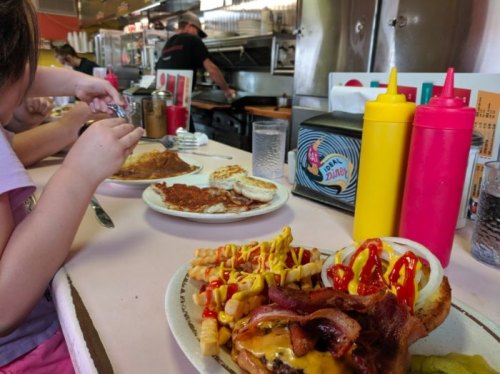 A diner table with a plate of a loaded burger, fries, and condiments, while someone prepares food in the background.