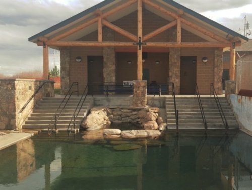 A rustic building with a stone facade and wooden beams, surrounded by water and steps leading down to a pond.