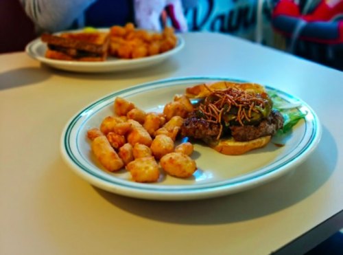 A plate with a burger topped with pickles and crispy onions, served with a side of golden tater tots.