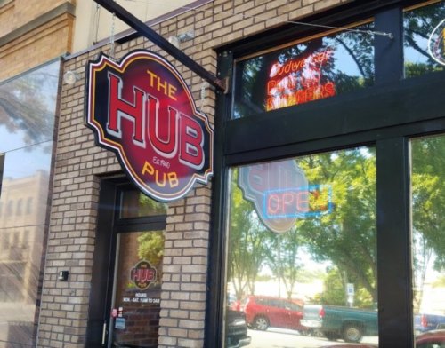 Sign for "The Hub Pub" with neon "OPEN" in the window, set against a brick building and trees outside.