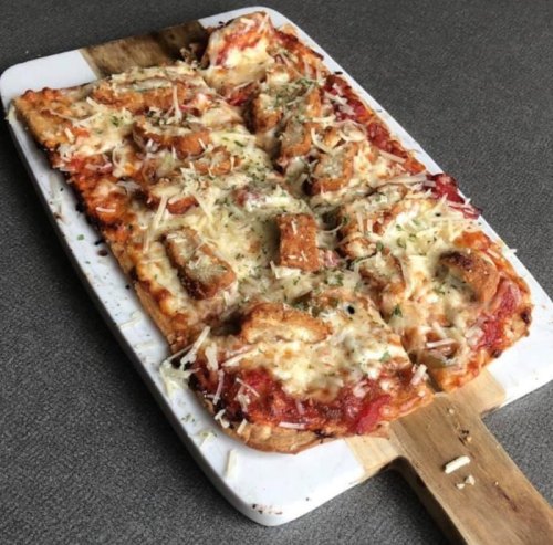 A rectangular pizza topped with cheese, sauce, and pieces of bread, served on a wooden cutting board.
