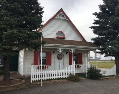 A charming white house with red shutters and a triangular roof, surrounded by green trees and a white picket fence.