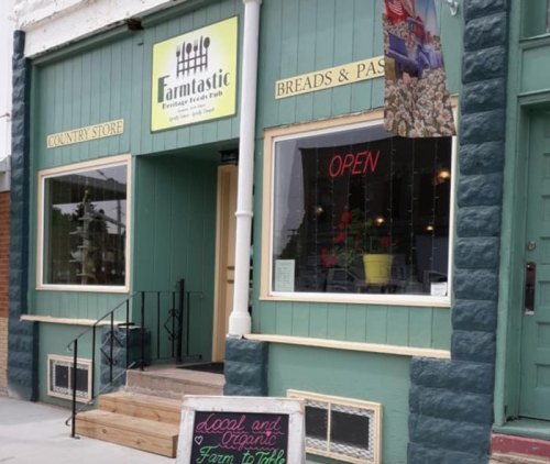 A green country store with a sign reading "Farmtastic," featuring an open sign and a chalkboard advertising local goods.