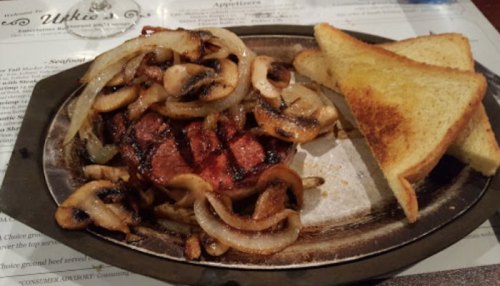 A sizzling steak topped with sautéed onions and mushrooms, served with two slices of toasted bread on a metal plate.