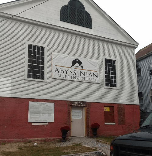 Exterior of the Abyssinian Meeting House, featuring a large sign and a red brick base with windows above.