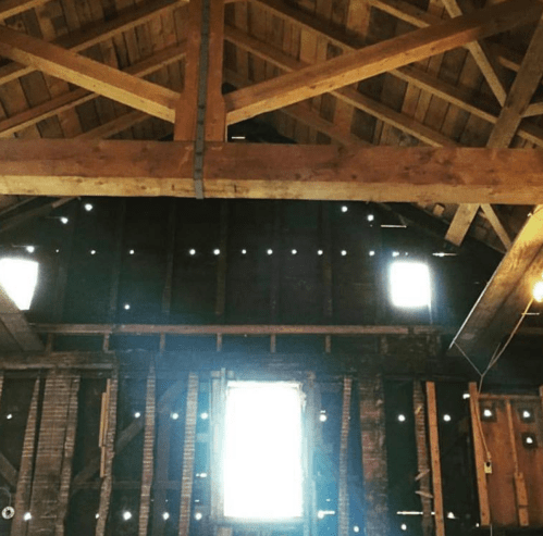 Interior of a rustic barn with wooden beams and a window, light streaming through gaps in the walls.