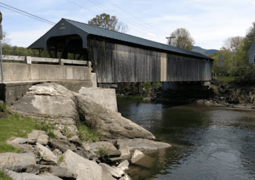 A wooden covered bridge spans a calm river, surrounded by greenery and a clear blue sky.
