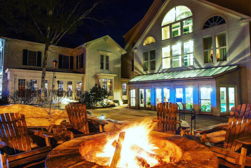 A cozy outdoor fire pit surrounded by wooden chairs, with a well-lit building and snow in the background at night.